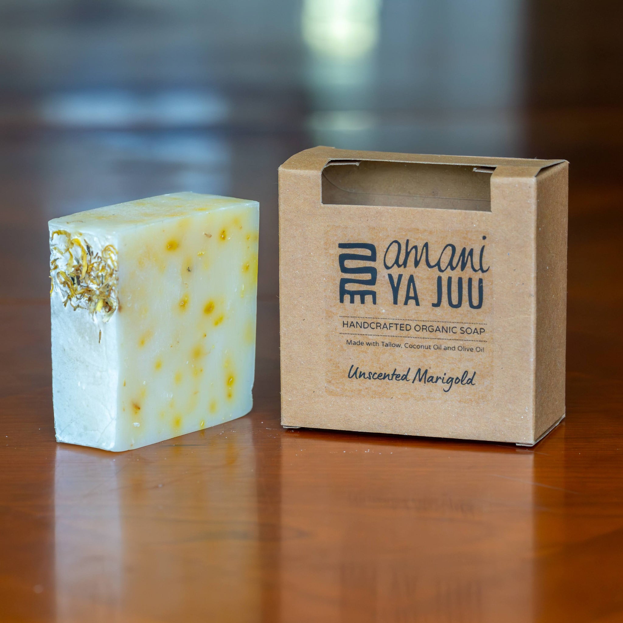 A handmade organic soap bar with yellow speckles and a gold foil accent stands next to its kraft paper packaging box on a polished wooden surface. The box is labeled 'amani YA JUU' and 'Handcrafted Organic Soap' with 'Unscented Marigold' written at the bottom.
