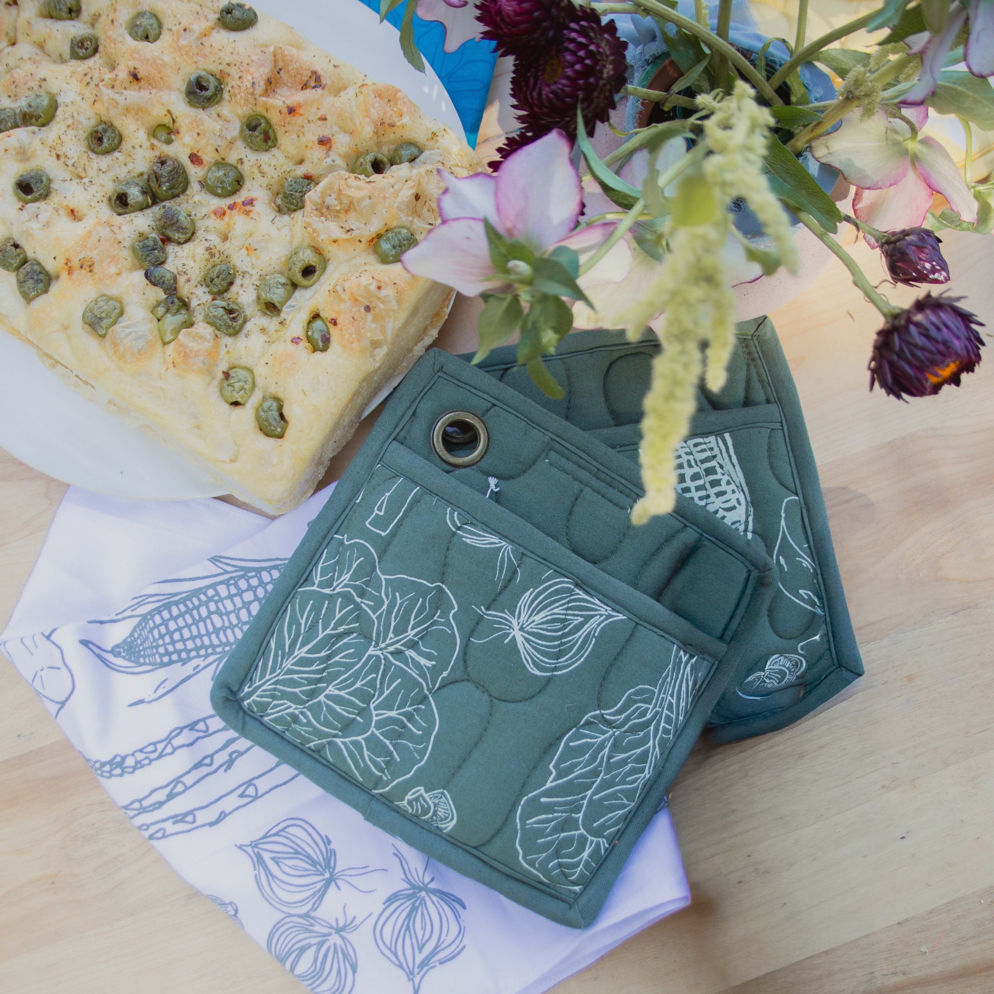 Green pot holders with leaf pattern on a wooden table with a plate of food and flowers.