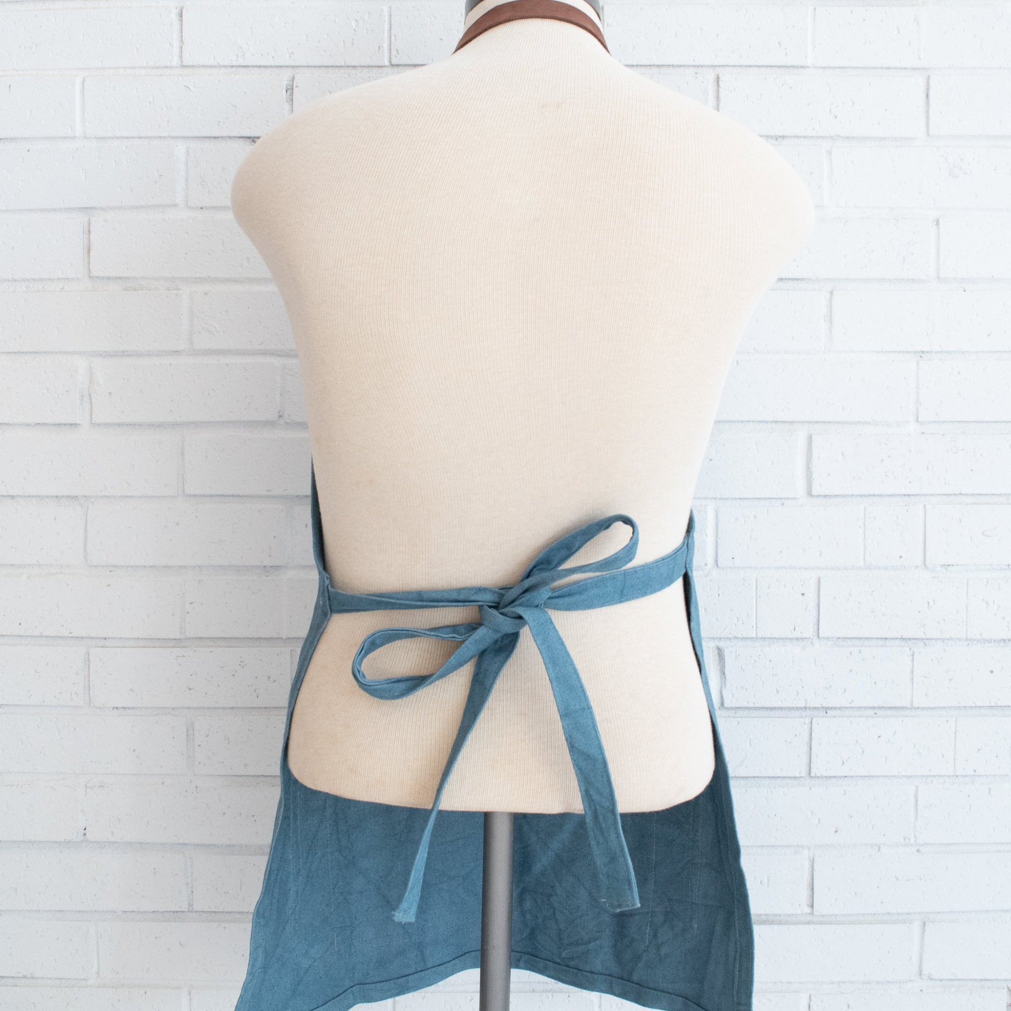 A blue apron tied around the waist of a cream-colored dress form mannequin, set against a white painted brick wall background.