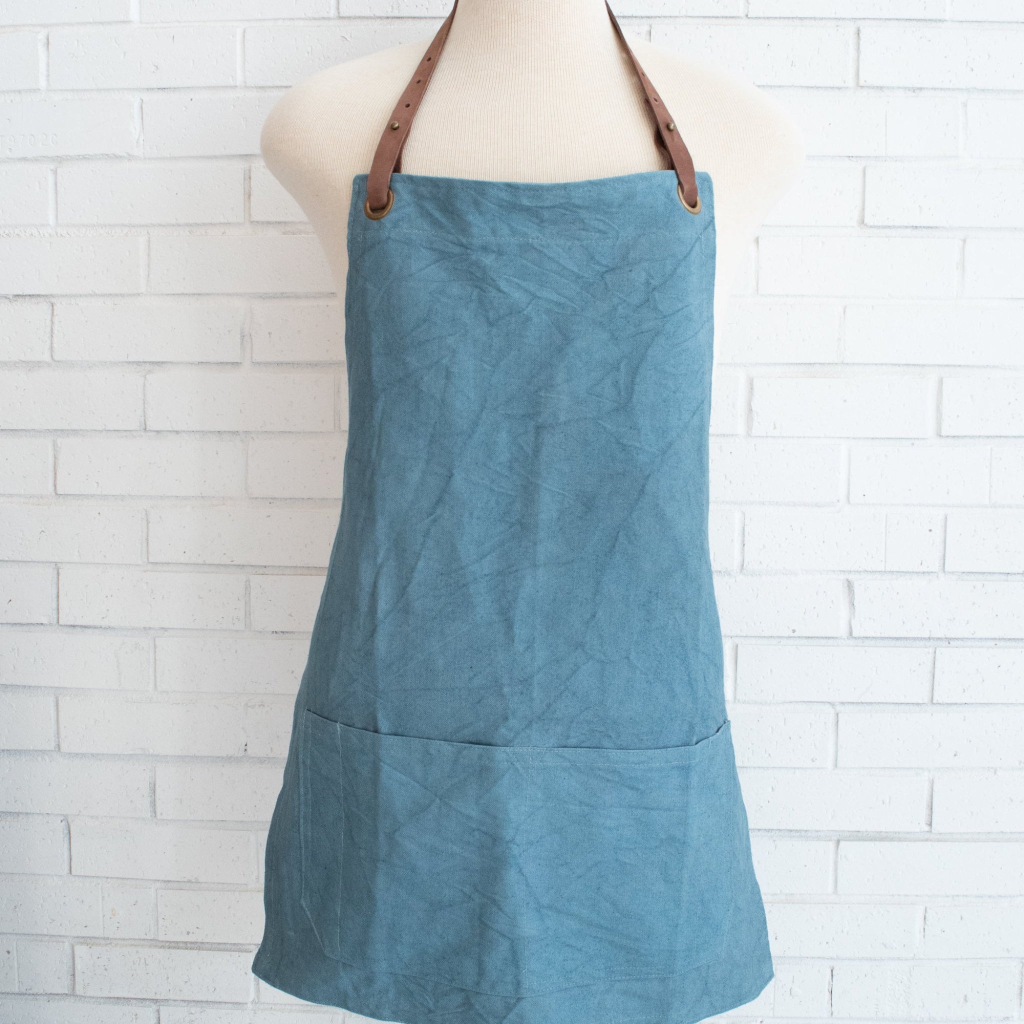 A dusty blue canvas apron with brown leather straps, displayed on a cream mannequin torso against a white brick wall background.