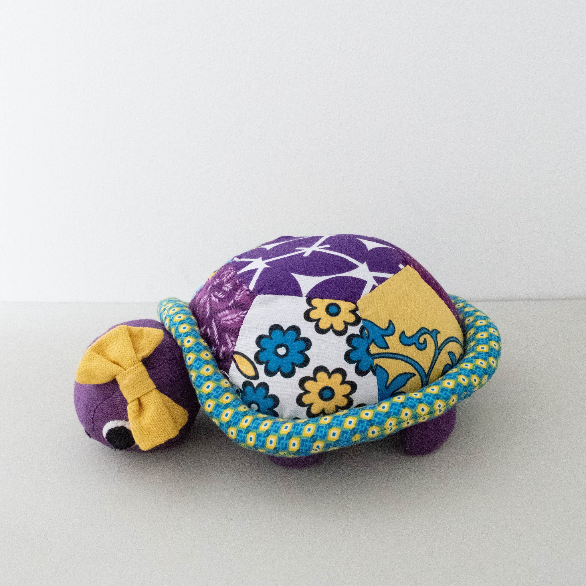 A handmade fabric turtle plush toy with a purple body, a patterned shell featuring purple, white, blue, and yellow floral designs, a blue and yellow patterned shell border, and a yellow bow on its head, set against a plain white background.