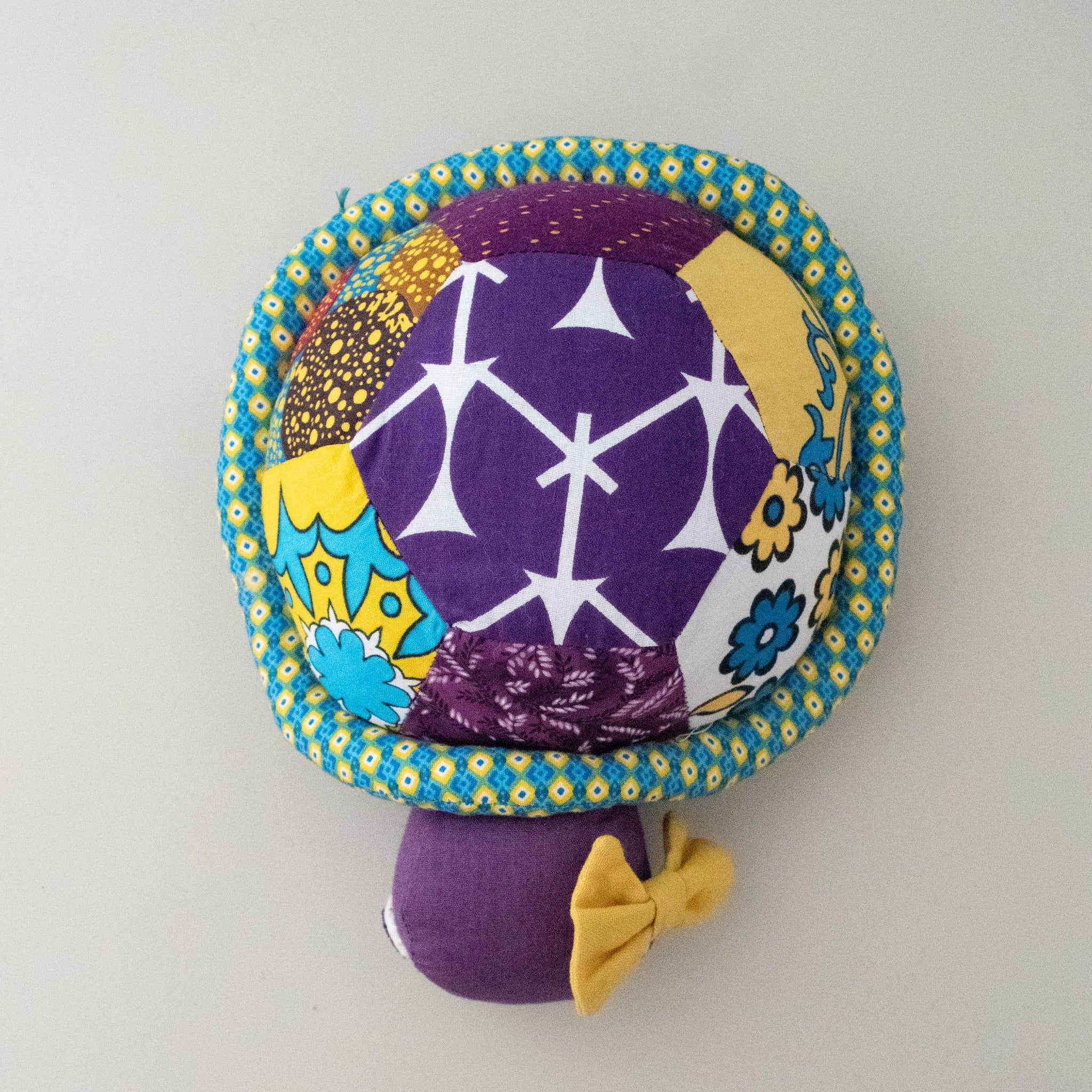 A handmade plush turtle made from colorful patterned fabrics, featuring a purple cap with white starburst motifs, a yellow and blue floral patterned border, and a purple stem with a small yellow bow, set against a plain light gray background.