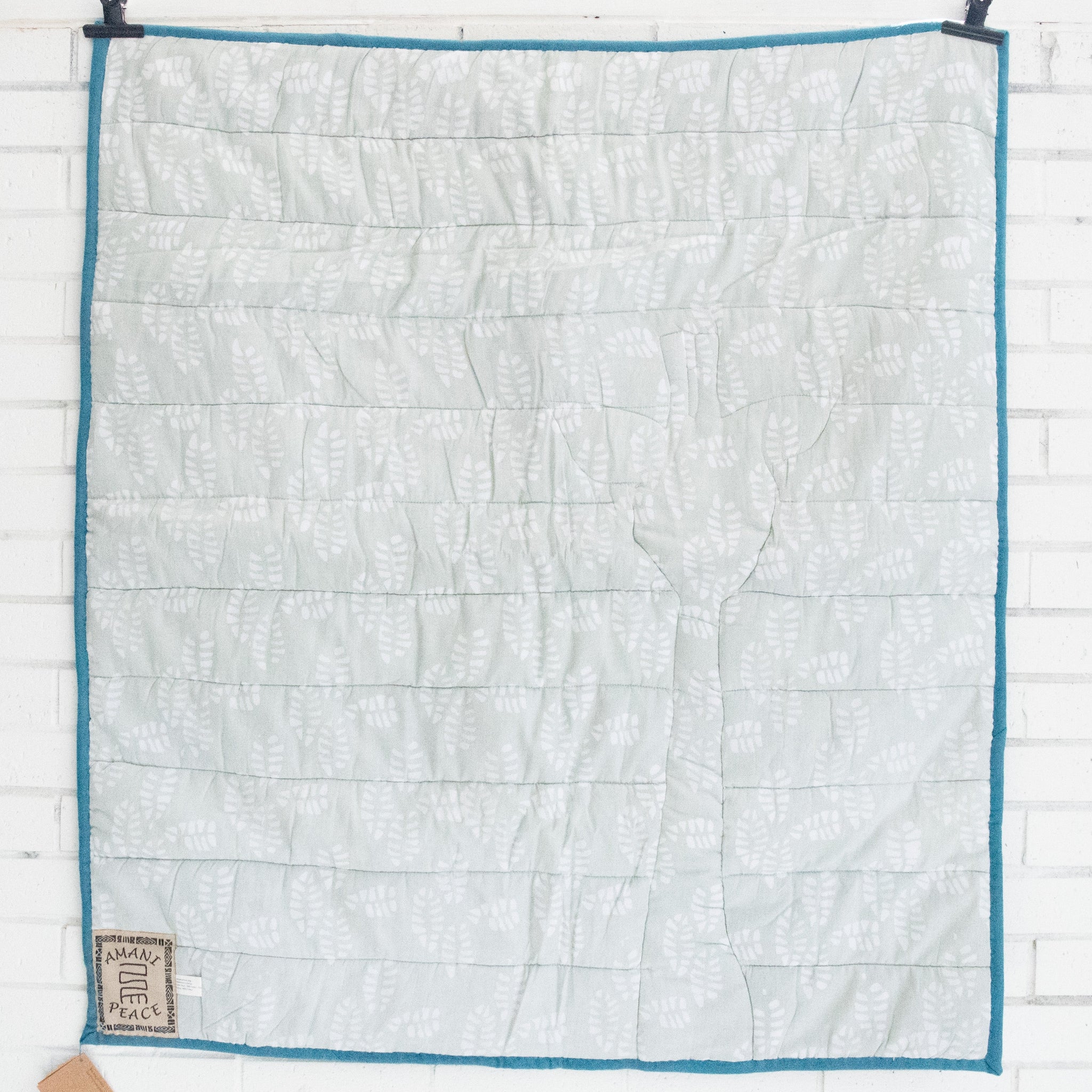 A square quilted blanket with a white leaf pattern on a light sage green background, finished with a teal blue border. 
