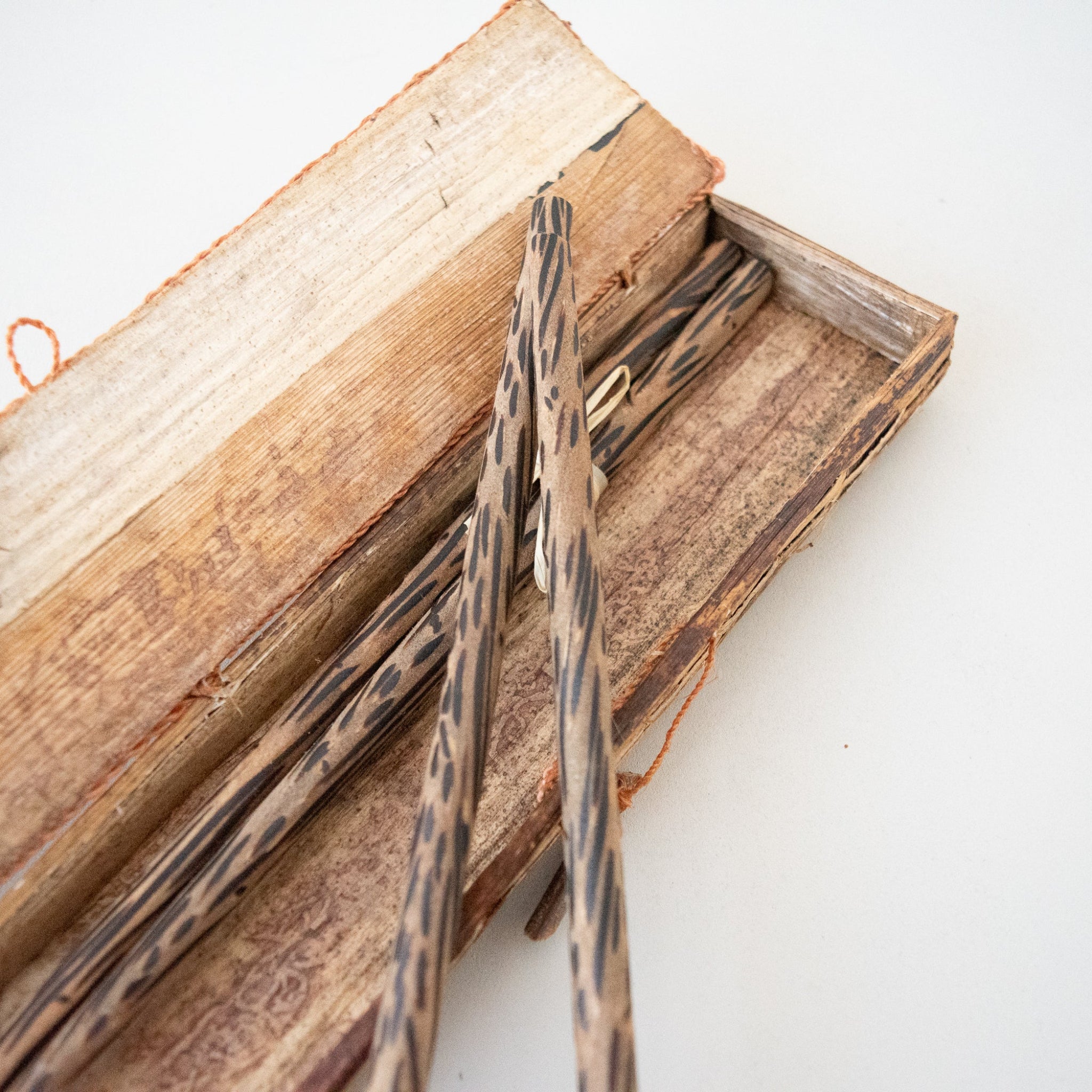 A close-up of a wooden box containing several long, narrow wooden chopsticks with dark grain patterns, photographed on a plain white background.