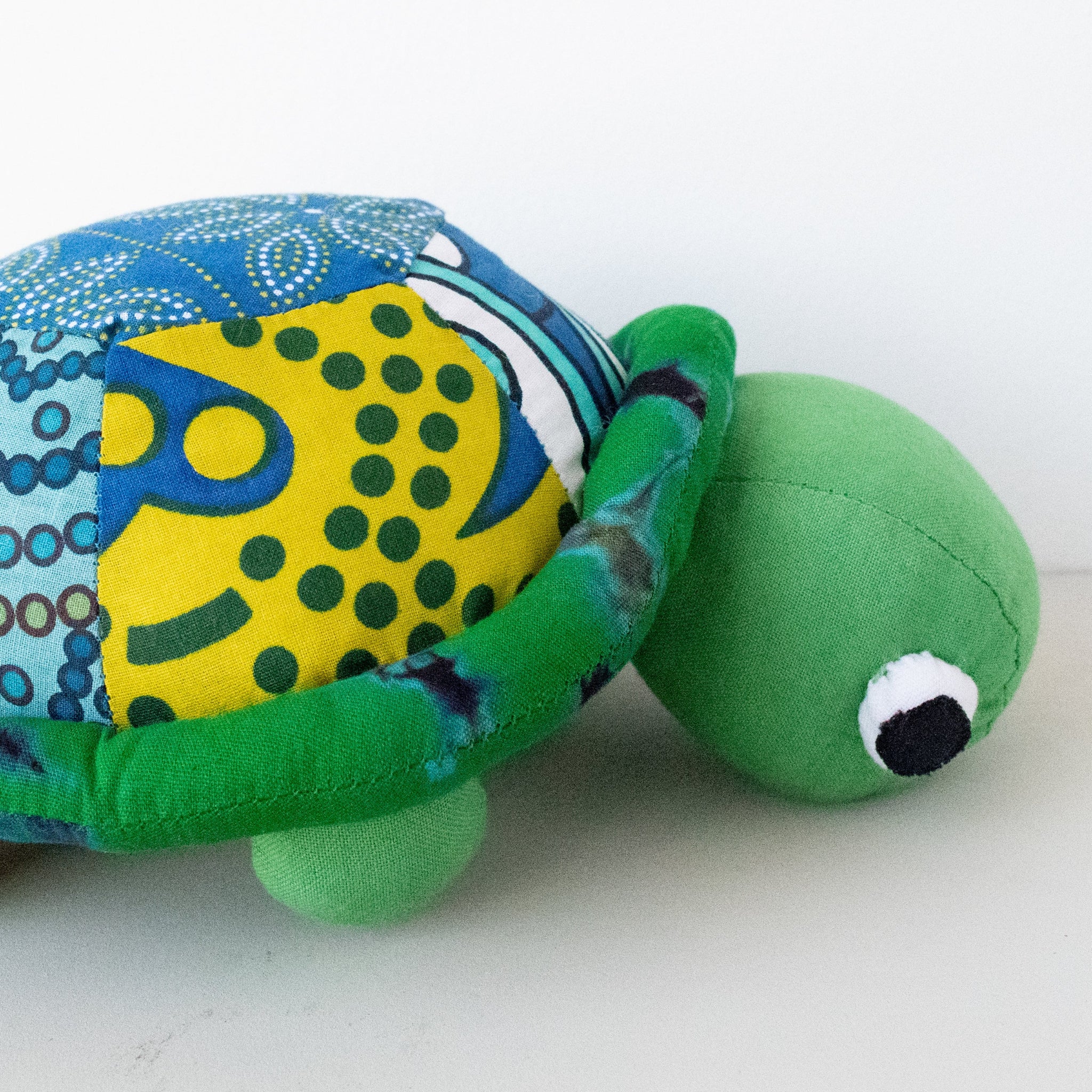 A handmade patchwork turtle plush toy with a green fabric head and legs, a patterned blue and yellow shell, and embroidered eyes, shown on a plain white background.