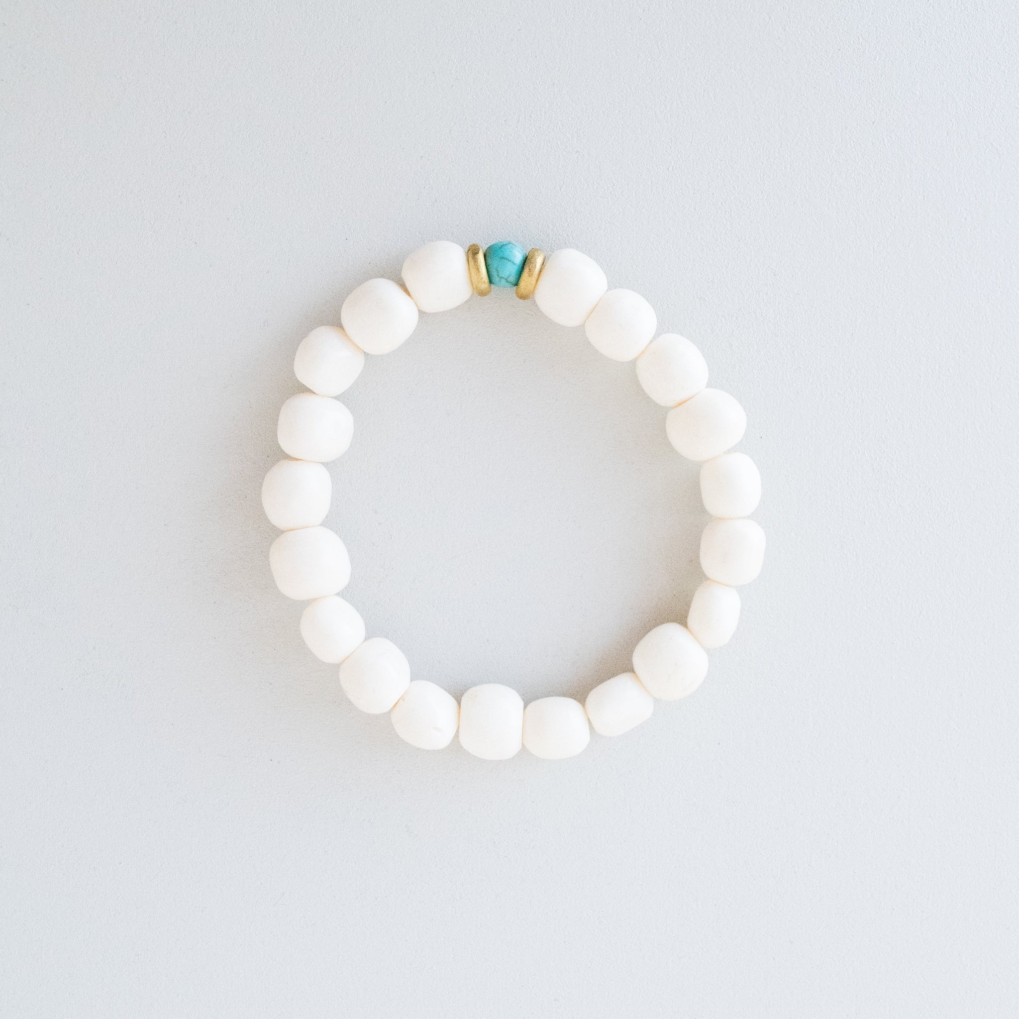 A beaded stretch bracelet made of matte white round beads, featuring a small turquoise stone and gold spacer at the top, displayed on a light gray background.