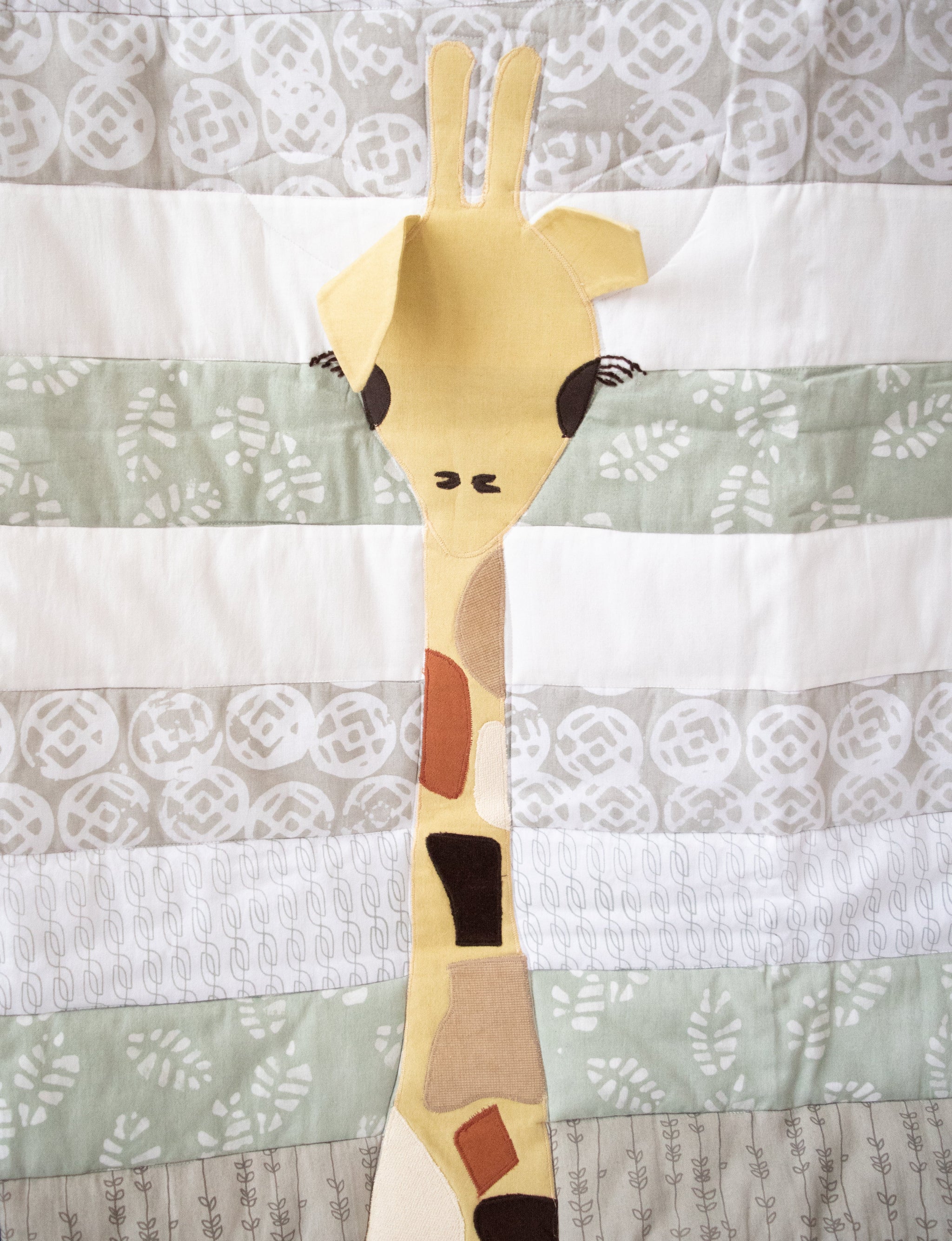 A close-up of a giraffe applique patch on a striped and patterned quilt. The patch is yellow with brown, beige, and black spots, set against a background of white, gray, and sage green patterned fabric bands.