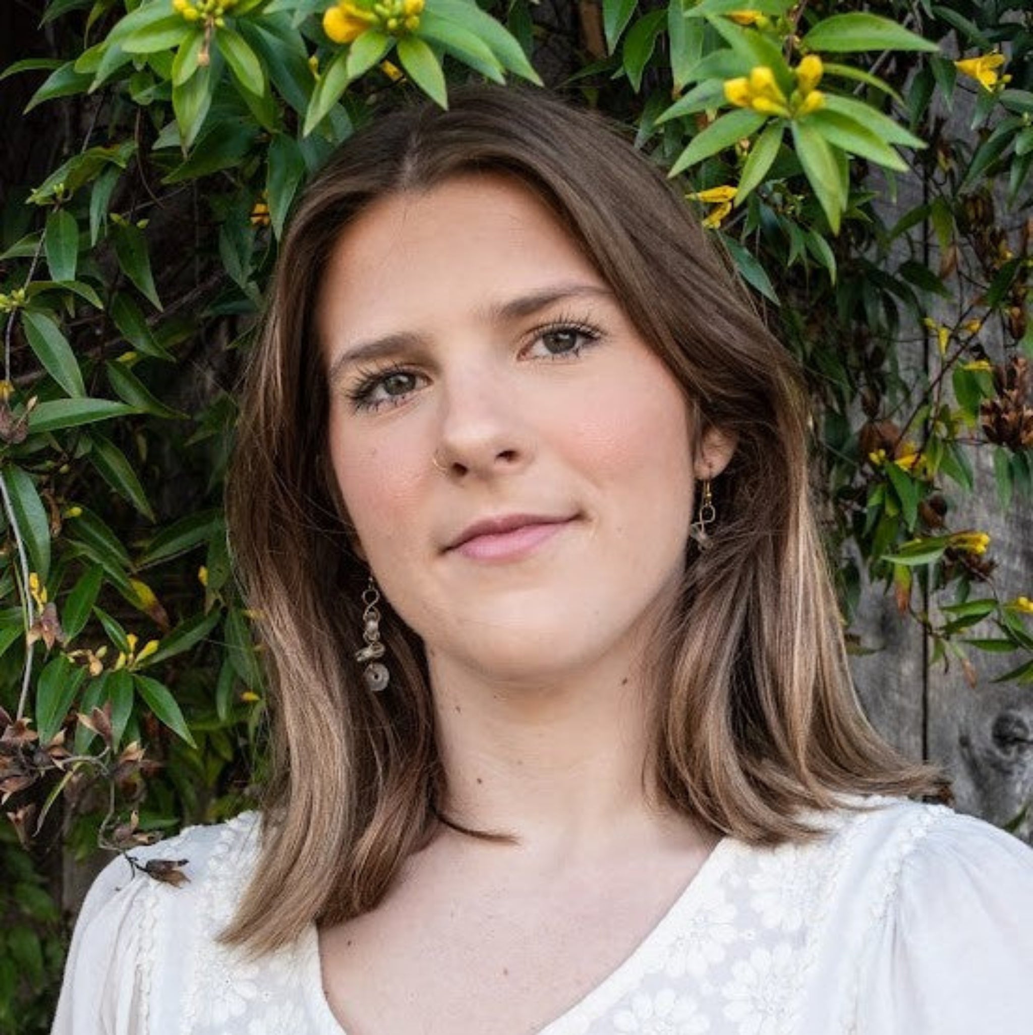Woman with shoulder-length brown hair standing in front of green foliage with yellow flowers.