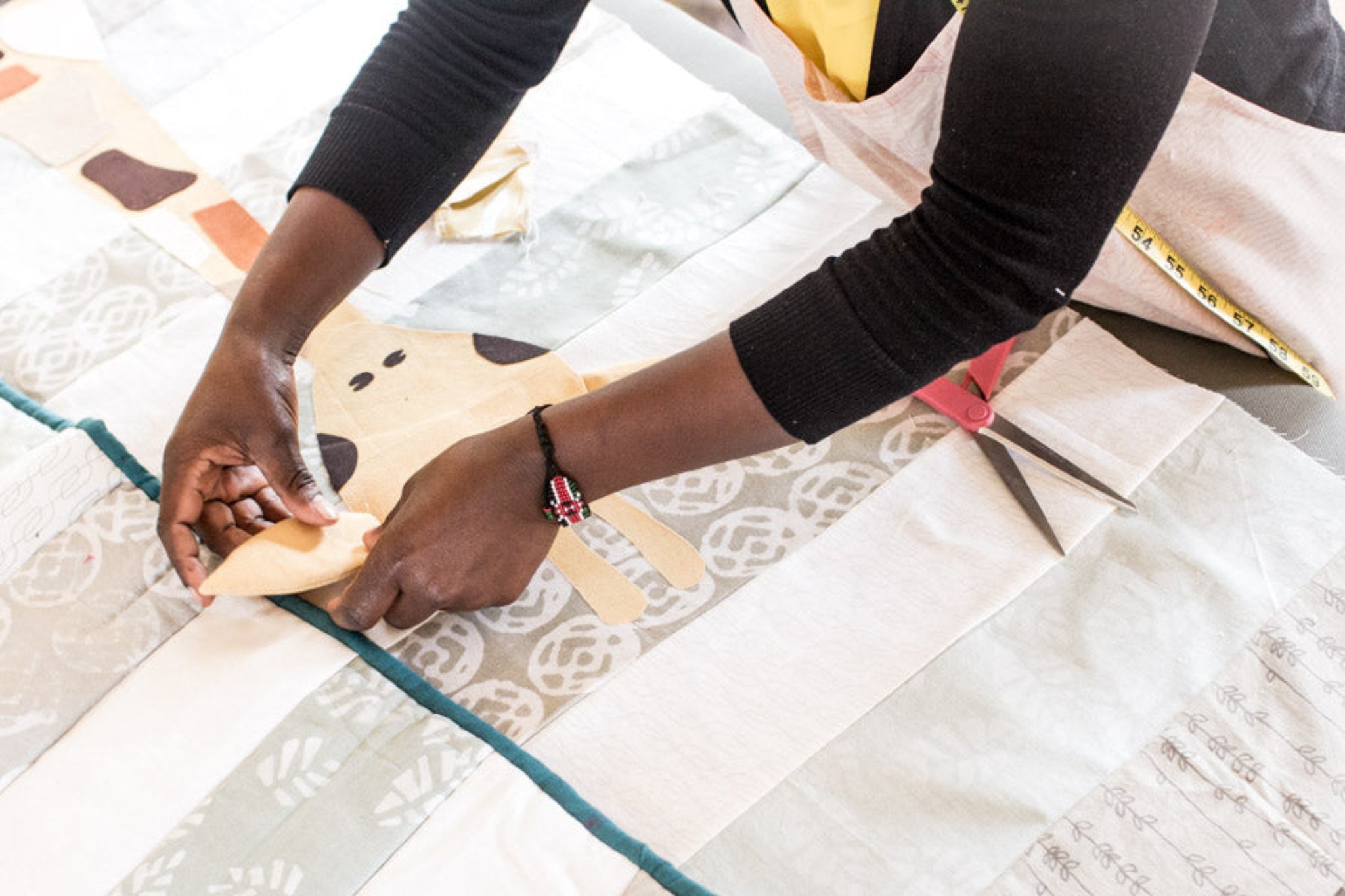A close-up of a person's hands placing a light beige fabric patch onto a large quilted textile with white, gray, and teal sections. The person is wearing a black long-sleeve top and a light apron, with a pair of scissors visible on the quilt.