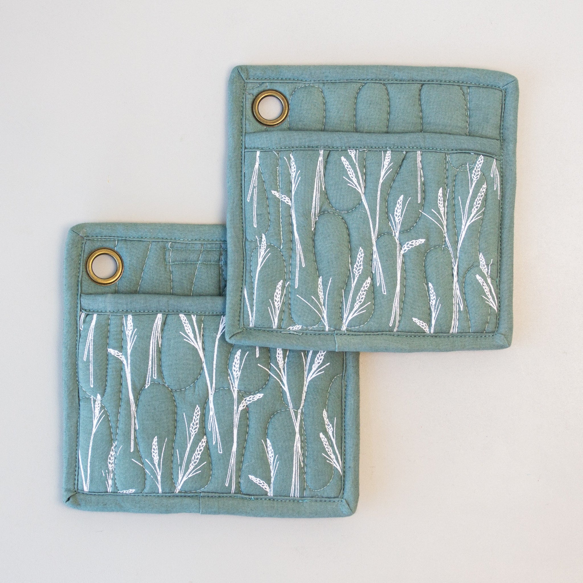 Two sage square pot holders with white wheat screen print and a pocket on each, with metal loops for hanging