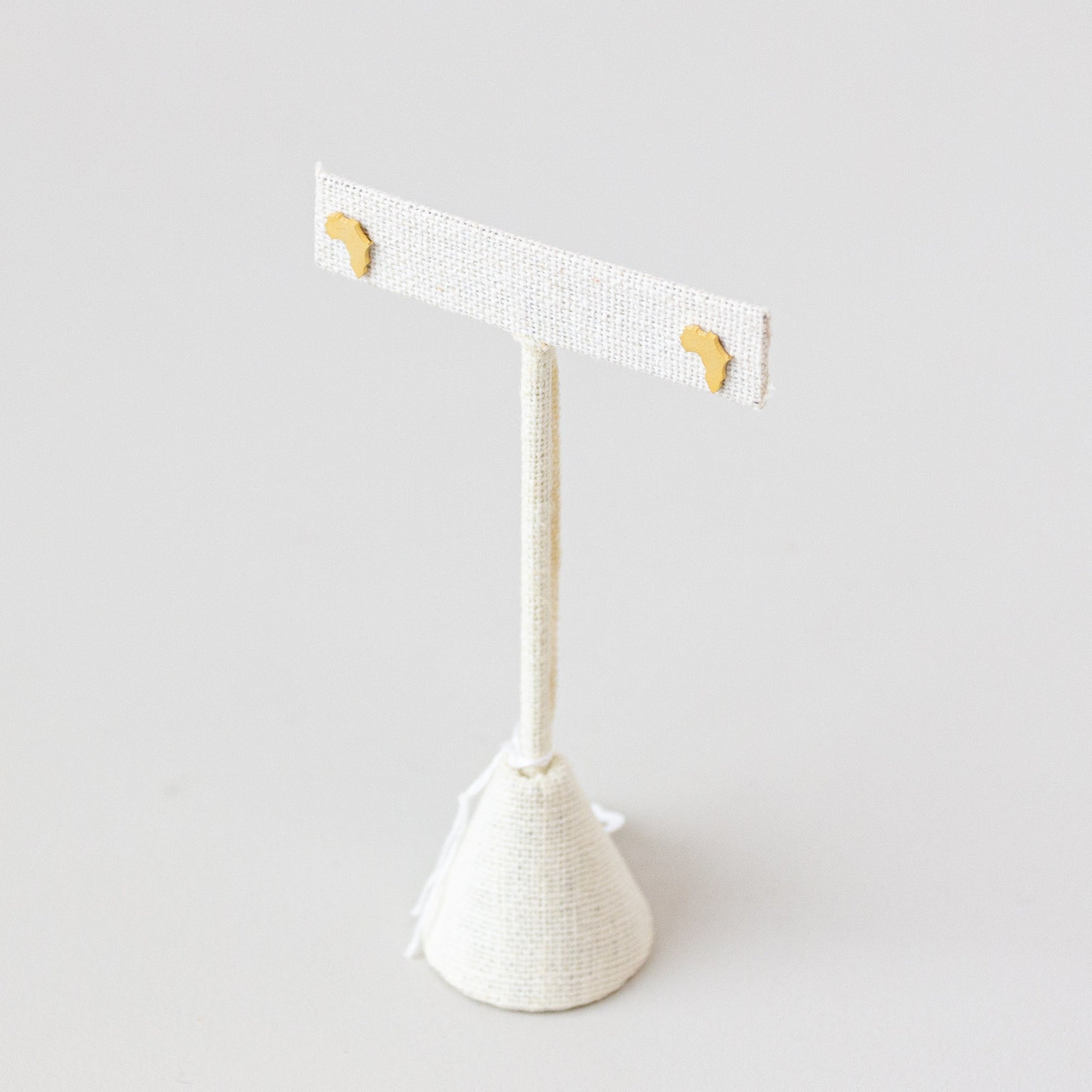 Brass earring studs shaped like the continent of Africa displayed on a white stand with a white background.