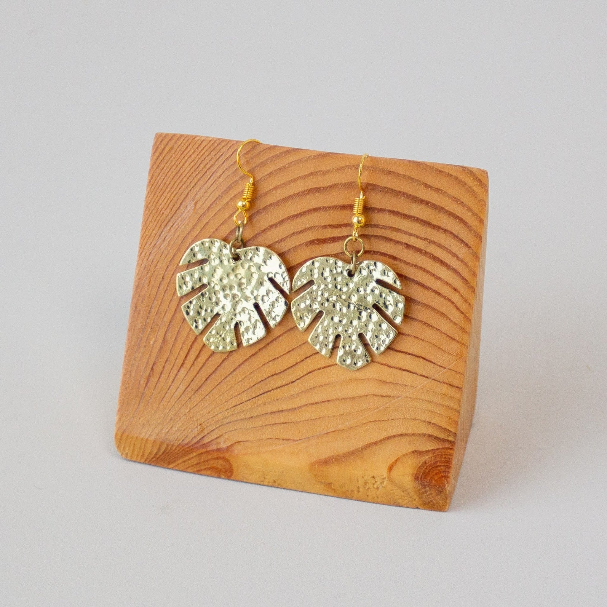Brass monstera leaf drop earrings displayed on wooden stand.
