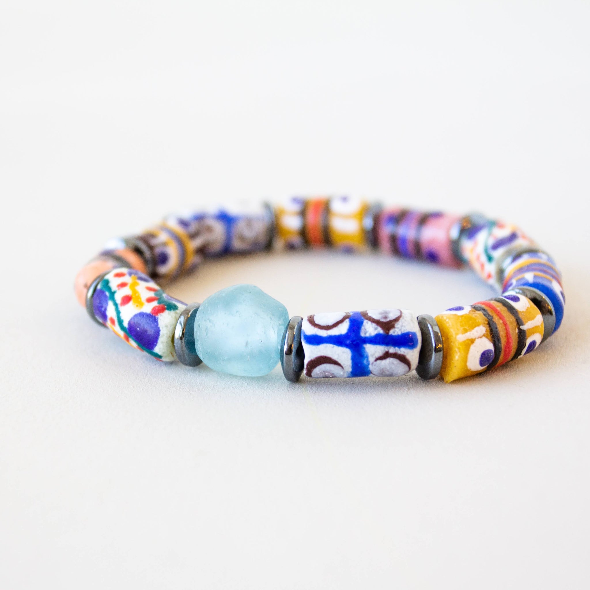 A handmade beaded stretch bracelet featuring a mix of colorful patterned glass beads, a light blue translucent bead, and one larger white bead with a blue cross, set against a plain light background.