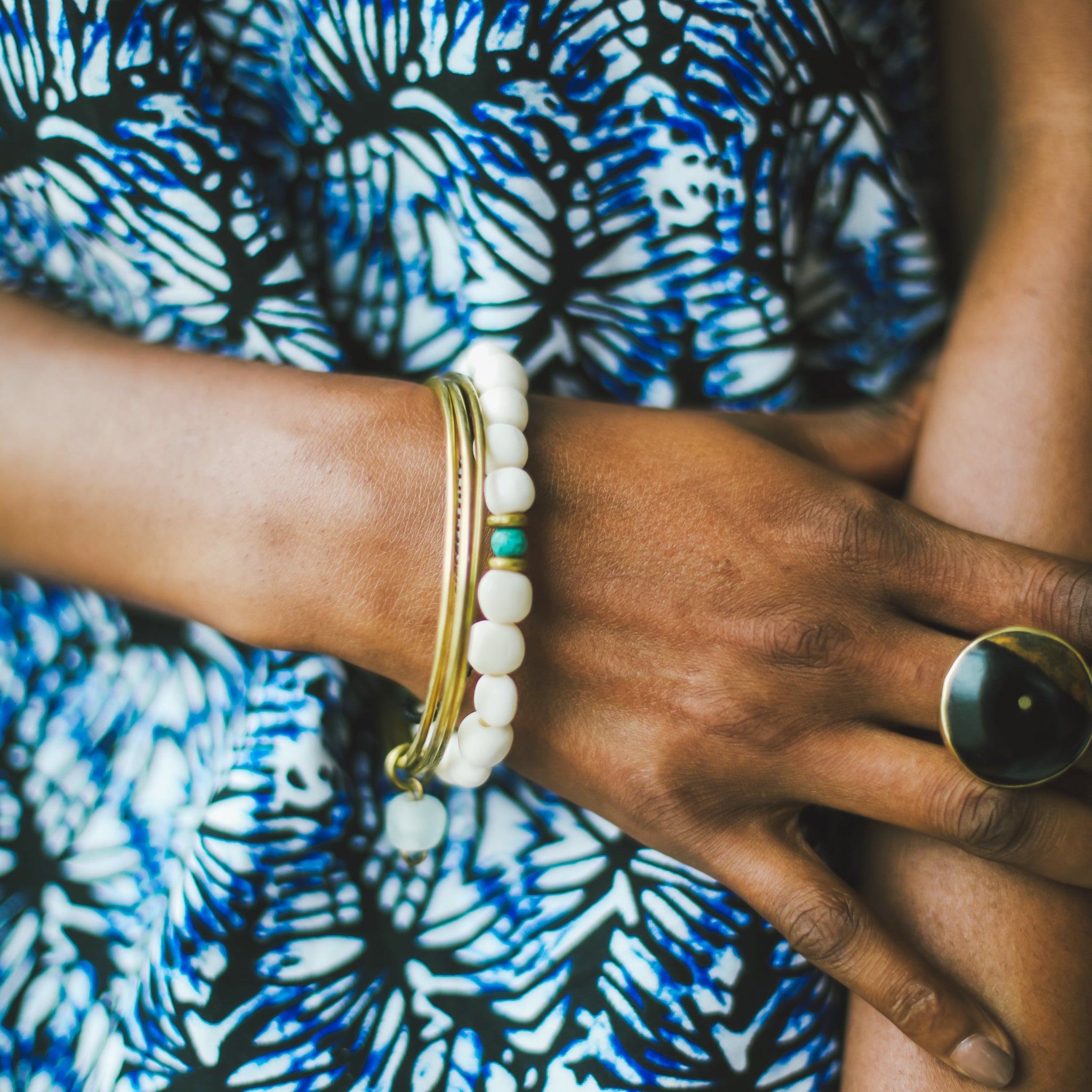 A close-up of a person's wrist adorned with gold bangles, a white beaded bracelet, and a small turquoise accent, with a large black ring on the finger. The background shows a blue and white patterned fabric.