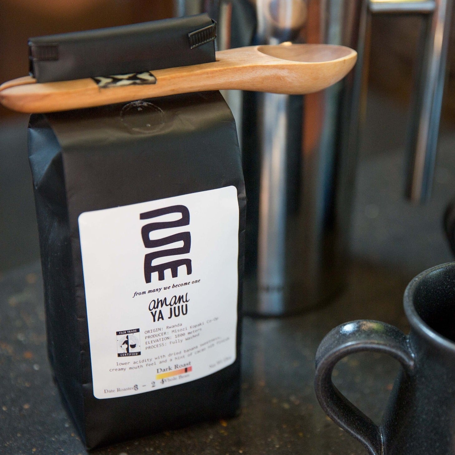 Black coffee bag with a white label and wooden spoon clip on counter with mugs