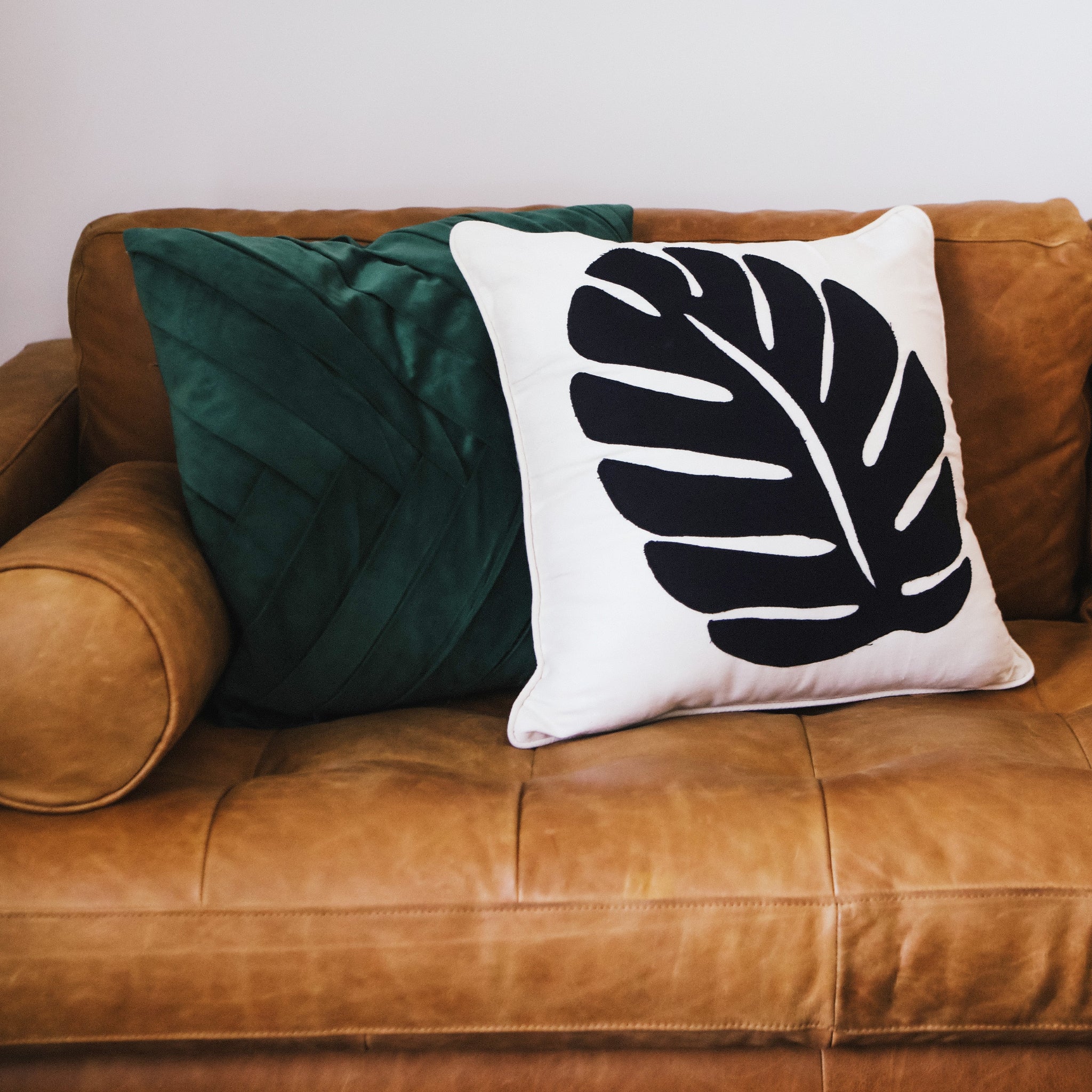 Two decorative pillows on a brown leather couch, one green and one with a leaf design.