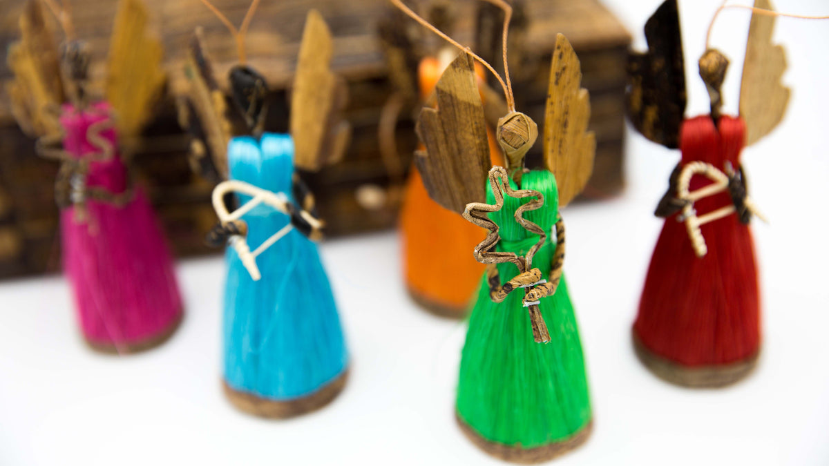 A set of small handmade angel ornaments with colorful bodies, wooden wings, and hanging strings, shown on a white background with a blurred wooden box behind them.