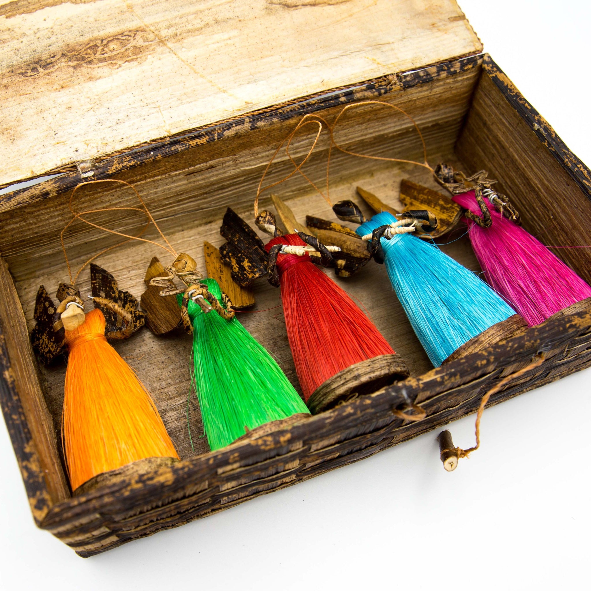 A set of colorful tassel ornaments shaped like angels, displayed inside a rustic wooden box. The ornaments feature vibrant tassels in orange, green, red, light blue, and magenta, with dark brown wings and wooden heads.