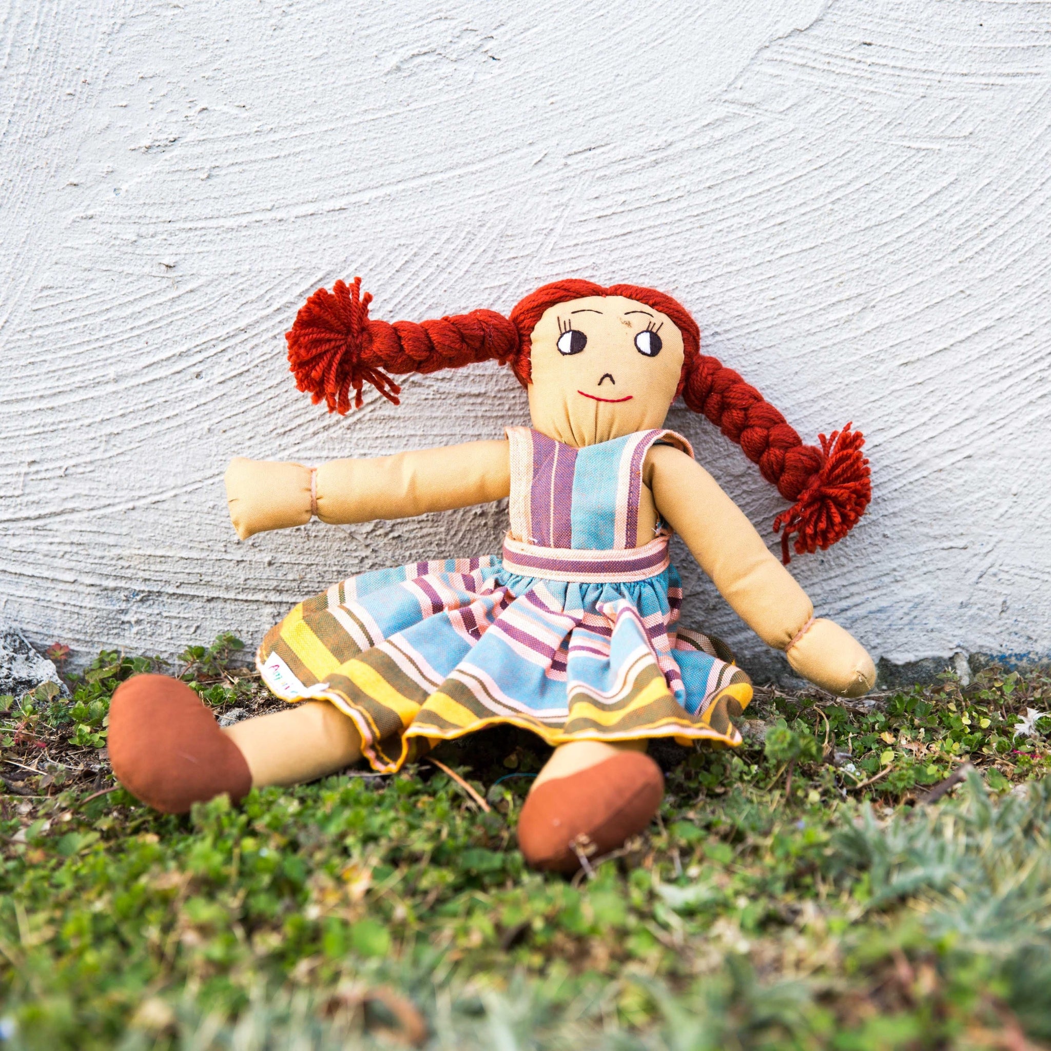Vintage-style doll with red hair and a colorful dress lying on grass against a textured wall.